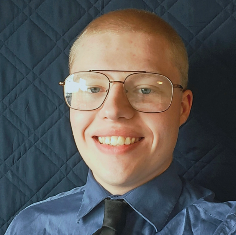 A young man with light skin, short blond hair, blue eyes, and brown aviator eyeglasses.
